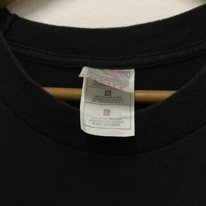 Peut inclure: Une &eacute;tiquette de t-shirt noire avec du texte blanc qui indique "Field of the Loom" et "100% coton pr&eacute;-r&eacute;tr&eacute;ci assembl&eacute; au Salvador avec des composants des &Eacute;tats-Unis."