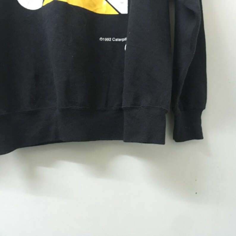May include: Black sweatshirt with a yellow and white graphic design. The graphic features a cartoon character and the text "&copy;1992 Caterpillar".