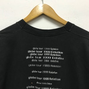 Peut inclure: T-shirt noir avec le texte "globe tour 1999 Relation" r&eacute;p&eacute;t&eacute; plusieurs fois dans un design empil&eacute;.