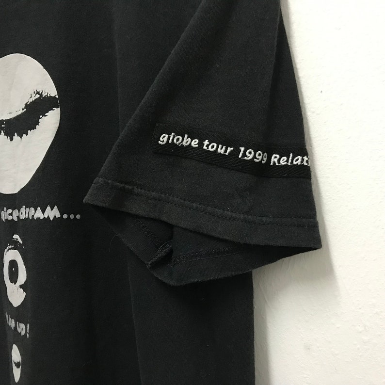 Peut inclure: T-shirt noir avec un imprim&eacute; graphique blanc d'un cercle avec un motif noir &agrave; l'int&eacute;rieur. Le texte "nicedream..." est imprim&eacute; sous le cercle. Le texte "globe tour 1999 Relax" est imprim&eacute; sur la manche du t-shirt.