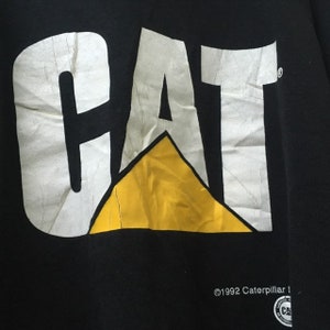 May include: Black t-shirt with a white "CAT" logo and a yellow triangle. The text "&copy;1992 Caterpillar Inc." is printed below the logo.
