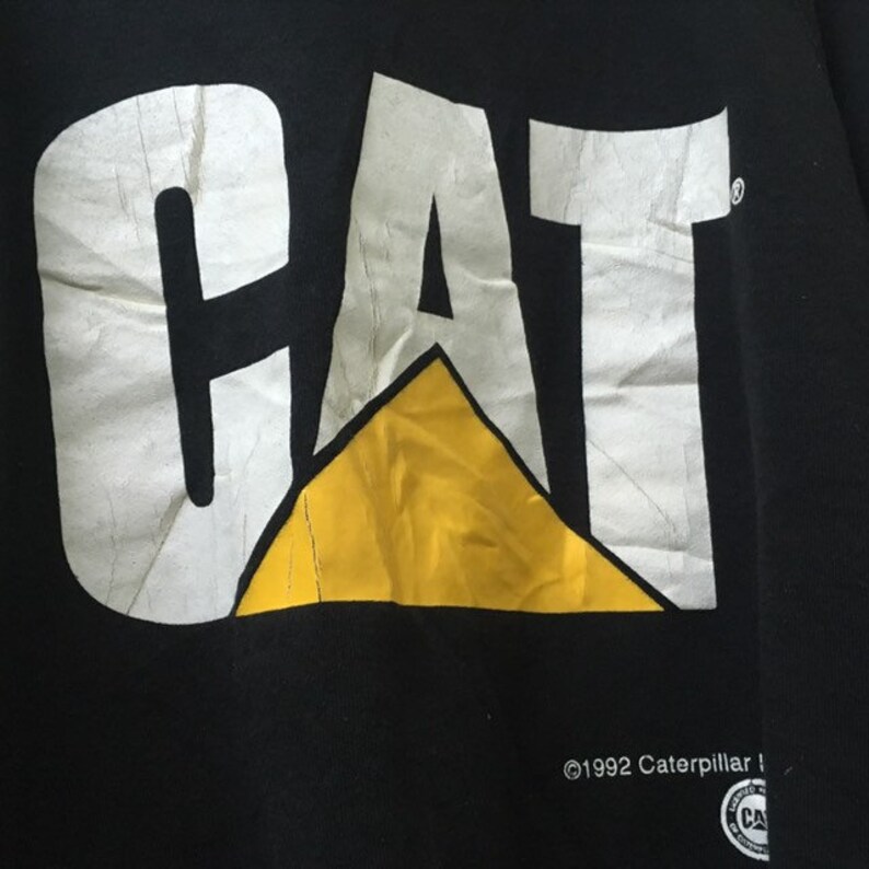 May include: Black t-shirt with a white "CAT" logo and a yellow triangle. The text "&copy;1992 Caterpillar Inc." is printed below the logo.