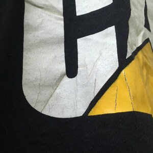 May include: Close-up of a black t-shirt with a white and yellow graphic design. The graphic features a stylized letter "H" with a yellow triangle.