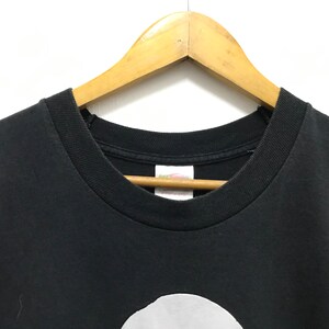 Peut inclure: T-shirt noir avec un cercle blanc sur le devant. Le t-shirt est suspendu &agrave; un cintre en bois.