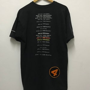 Peut inclure: T-shirt noir avec un grand chiffre 4 orange en bas &agrave; droite. Le dos du t-shirt porte le texte "globe tour 1999 Relation" r&eacute;p&eacute;t&eacute; plusieurs fois dans diff&eacute;rentes couleurs et polices.
