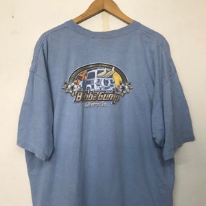 Könnte beinhalten: Ein hellblaues T-Shirt mit einem Aufdruck eines Oldtimers mit Surfbrettern auf dem Dach. Der Aufdruck ist von einem gelben Kreis umgeben, in dem der Text "Bubba Gump Shrimp Co." in brauner Schrift steht.