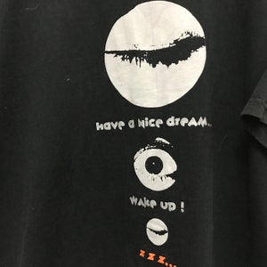 Peut inclure: T-shirt noir avec un cercle blanc contenant un motif noir, le texte "have a nice dream", un &oelig;il blanc avec une pupille noire, le texte "wake up!" et un croissant de lune blanc avec un motif noir. Le texte "zzz..." est sous la lune.