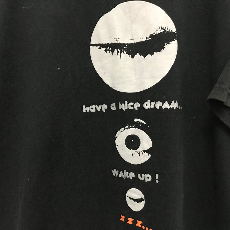 Peut inclure: T-shirt noir avec un cercle blanc contenant un motif noir, le texte "have a nice dream", un &oelig;il blanc avec une pupille noire, le texte "wake up!" et un croissant de lune blanc avec un motif noir. Le texte "zzz..." est sous la lune.