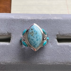 Könnte beinhalten: Ein silberner Ring mit einem großen, hellblauen Edelstein, der von kleineren Türkis-Steinen umgeben ist.