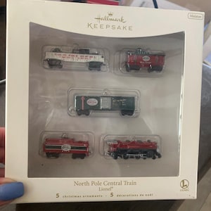 以下が含まれることがあります： Hallmark Keepsakeの、5つのミニチュアクリスマス列車のオーナメントのボックスセット。セットには、「North Pole Central Rail」のテキストが入った白い車両、赤いカブース、緑の貨車、2つの赤い機関車が含まれています。箱には「North Pole Central Train」と書かれています。