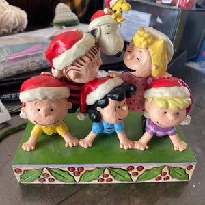 Puede incluir: Una figura navideña con personajes de Peanuts, incluyendo a Charlie Brown, Snoopy, Lucy, Linus y Sally, todos con gorros de Papá Noel. La base es verde con detalles de acebo y bayas. Un artículo de colección.