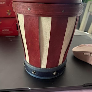 Può includere: Un cesto decorativo a tema patriottico con coperchio. Il cesto presenta strisce verticali rosse e bianche alternate, una fascia blu scuro con dettagli a stella e un coperchio rosso scuro. Il cesto è appoggiato su una superficie nera.