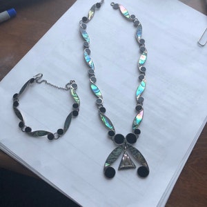 May include: A silver necklace and bracelet set with black and iridescent abalone shell accents. The necklace features a pendant with a triangular abalone shell in the center.