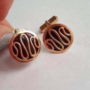 May include: A pair of gold-toned cufflinks with a circular design. Each cufflink features a dark red center with a gold-colored, swirling pattern. The cufflinks are shown on a white background.