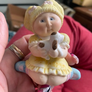 Puede incluir: Una figurita de porcelana de una muñeca Cabbage Patch Kid con cabello rubio en coletas, vestida con un vestido amarillo y zapatos azules, sosteniendo un cachorro blanco y marrón.