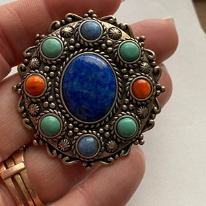May include: A vintage brooch with a silver-tone metal frame. The brooch features a large, oval, deep blue stone in the centre, surrounded by smaller blue, turquoise, and orange stones. The design is ornate and detailed, with a classic, timeless appeal.