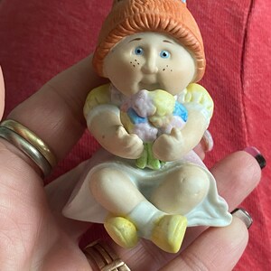 May include: A porcelain figurine of a Cabbage Patch Kid doll with red hair, a blue bow, and a pink dress. The doll is holding a bouquet of colorful flowers.