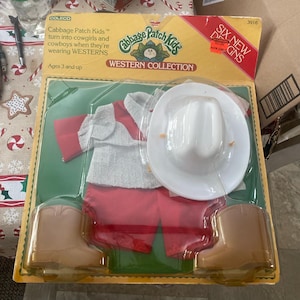 May include: A Cabbage Patch Kids Western Collection toy set. The set includes a white cowboy hat, red trousers, a grey and red shirt, and brown cowboy boots. The packaging is yellow and green.