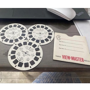 May include: Three white View-Master reels with black rectangular viewing windows and text. A View-Master reel sleeve is next to the reels. The sleeve has the words "VIEW-MASTER REELS" in red and white text.