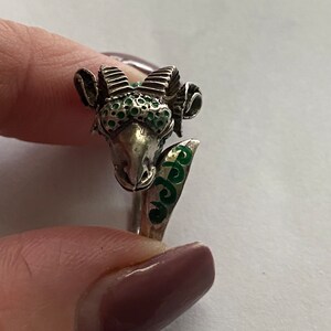 May include: Silver ring featuring a detailed ram's head design. The ram's horns and face are decorated with green enamel.
