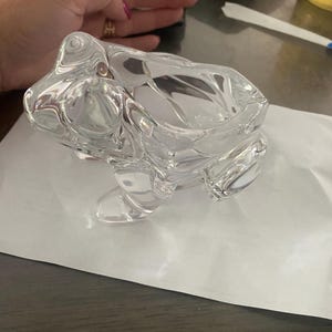 以下が含まれることがあります： 詳細な特徴を持つ透明なガラス製のカエルの置物。カエルは白い表面に置かれています。置物は滑らかで反射する表面を持ち、デザインはカエルの本質を捉えています。