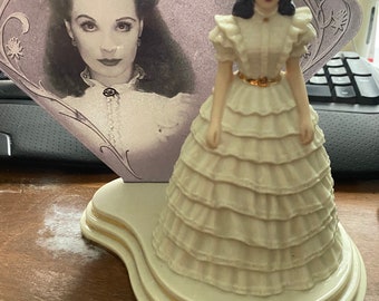 Peut inclure: Une figurine en porcelaine d'une femme en robe blanche avec une ceinture dorée. La figurine est sur un socle à trois niveaux. La figurine est devant une photo en noir et blanc d'une femme.
