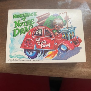Könnte beinhalten: Eine Vintage-Sammelkarte mit einer Cartoon-Illustration eines roten Autos, das von einem buckligen Charakter gefahren wird. Das Auto trägt den Text "Lil' Ding Dong" und "Hooker Headers". Die Karte stammt aus der Serie 1980 Topps Chewing Gum, Inc.