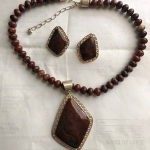 May include: A red and brown stone necklace with a matching pendant and earrings. The necklace is made of round beads and the pendant and earrings are in a diamond shape.