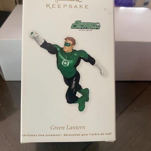 May include: A Hallmark Keepsake Christmas tree ornament featuring the Green Lantern superhero. The ornament depicts the Green Lantern in a dynamic pose, with a green and black costume. The box has the text "Green Lantern" and "christmas tree ornament".