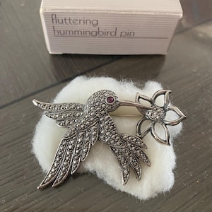 Op de afbeelding: Een zilveren kolibrie broche met een rode strass-oog en een bloemmotief. De broche ligt op een witte achtergrond en op de doos staat "fluttering hummingbird pin".