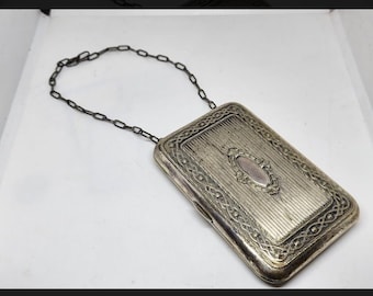 May include: A silver chain attached to a silver rectangular box with an ornate design. The box has a raised oval in the center with a decorative border.