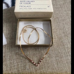 May include: A gold chain necklace with a V-shaped pendant of simulated ruby and rhinestone stones. The necklace is 16 inches long. The necklace is in a box with the text "Avon Galaxy of Color Necklace Simulated Ruby and Rhinestone 16" and "To Clean: Wash in mild, soapy water. Dry with a soft cloth."
