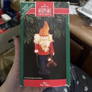 May include: Hallmark Keepsake ornament of Max The Tailor, a North Pole Nutcracker. The ornament features a red jacket, blue pants, and an orange hat. It holds a small brown teddy bear and a sewing tool. The box reads "Nutcracker Opens and Closes!"