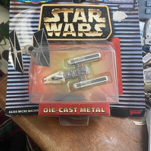 May include: A silver and yellow die-cast metal Star Wars X-Wing fighter toy in a plastic blister pack. The packaging features the words "Micro Machines" and "Star Wars" in bold letters. The warning label reads "CHOKING HAZARD - Small parts. Not for children under 3 years. Ages 4 and Up."