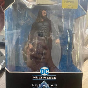 Könnte beinhalten: Verpackte Aquaman-Actionfigur aus der DC Multiverse-Serie. Die Figur befindet sich in einer transparenten Kunststoffhülle, mit dem Aquaman-Logo und dem Text "Aquaman and the Lost Kingdom" auf der Verpackung. Die Figur trägt ein schwarz-braunes Kostüm.