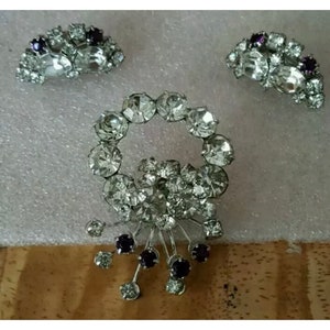 May include: A set of three vintage rhinestone jewelry pieces. The brooch features a swirl design with clear rhinestones and purple accents. The earrings are a matching set of clear rhinestones with purple accents.