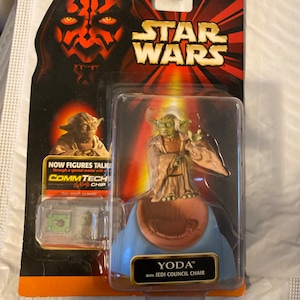 Könnte beinhalten: Eine Star Wars Yoda Actionfigur mit einem Jedi-Rat-Stuhl. Die Figur ist grün und trägt eine braune Robe. Der Stuhl ist blau und braun. Die Verpackung ist rot, schwarz und gelb mit dem Schriftzug "Star Wars". Die Figur ist Teil der CommTech-Linie, die es Figuren ermöglicht, durch einen speziellen Leser zu sprechen.