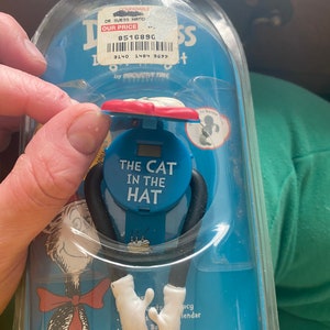 May include: A Dr. Seuss "The Cat in the Hat" watch in its original packaging. The watch features a blue face with the text "The Cat in the Hat". The watch has black arms and white gloved hands. The packaging is blue.
