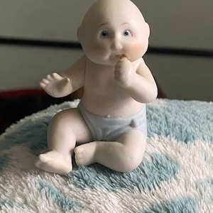 May include: A porcelain figurine of a baby sitting with its thumb in its mouth, wearing a blue nappy. The figurine is white with blue eyes and is sitting on a blue and white striped surface.