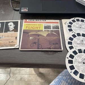 May include: Vintage View-Master set featuring "History of Flight" with 21 stereo pictures. Includes a box and several reels with images of early aircraft and aviation pioneers. The set is on a dark surface.