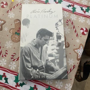 Peut inclure: Une couverture de livre en noir et blanc avec Elvis Presley. Le titre est "Elvis Presley Platinum" au-dessus d'une photo d'Elvis jouant du piano. Le texte "A Life in Music" est au bas de la couverture.