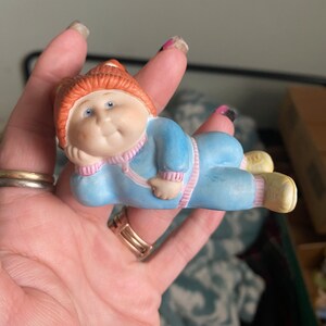 Könnte beinhalten: Eine Keramikfigur eines molligen Babys, das einen blauen und rosafarbenen Trainingsanzug mit einem braunen Hut trägt. Das Baby liegt auf der Seite und ruht seinen Kopf auf seiner Hand.