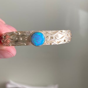 May include: A silver bracelet with a blue opal gemstone set in the center. The bracelet has a wavy design and is decorated with small dots.