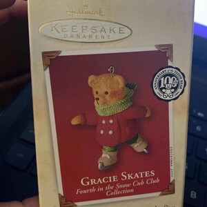 Peut inclure: Ornement Hallmark Keepsake représentant un ours en peluche nommé Gracie, portant un manteau rouge et une écharpe verte, faisant du patin à glace. La boîte indique "Gracie Skates" et "Fourth in the Snow Cub Club Collection". L'ornement célèbre le 100e anniversaire de l'ours en peluche.