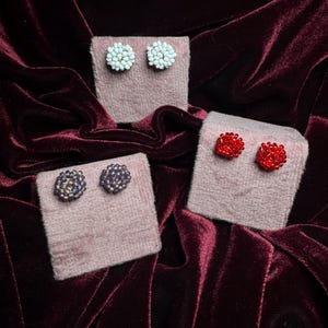 Könnte beinhalten: Drei Paar Perlenohrstecker in Weiß, Rot und Lila. Jedes Paar wird auf einem kleinen, rosa, rechteckigen Display präsentiert. Die Ohrringe sind in einem floralen Design angeordnet. Der Hintergrund ist ein dunkelburgunderfarbener Samt.