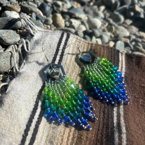 Pode incluir: Um par de brincos de contas com um gradiente de contas verdes e azuis. Os brincos têm um topo geométrico prateado com uma pequena pedra iridescente. As contas são dispostas em um padrão de franjas.