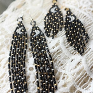 May include: Two pairs of long, dangling beaded earrings featuring black, gold, and silver seed beads. The earrings are constructed with multiple vertical strands of beads attached to a top section that displays a small white crescent moon motif.