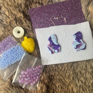 Könnte beinhalten: Eine Sammlung von Bastelmaterialien, darunter zwei abstrakte Ohrringe mit lila und blauen Mustern, Ohrringhaken, eine Spule weißen Fadens, eine gelbe fruchtförmige Perle und Beutel mit blauen und lila Perlen. Die Artikel sind auf einer strukturierten braunen Oberfläche angeordnet.