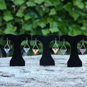 May include: Three pairs of triangle-shaped dangle earrings featuring intricate beadwork in blue, orange, and purple colour palettes. Each earring is suspended from a silver-toned metal hook and hangs from a black velvet display stand.