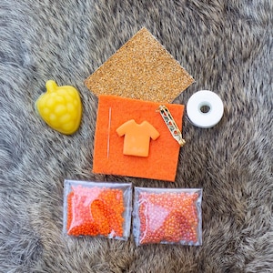 May include: A selection of craft materials, including orange beads in small bags, a yellow heart-shaped object, a gold glittery square, an orange felt square with a small orange shirt, a gold pin, and a roll of white tape. The items are on a textured, grey surface.